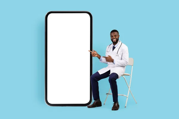远程医疗概念。友好的黑人医生坐在大空白智能手机旁边的椅子上 — 图库照片
