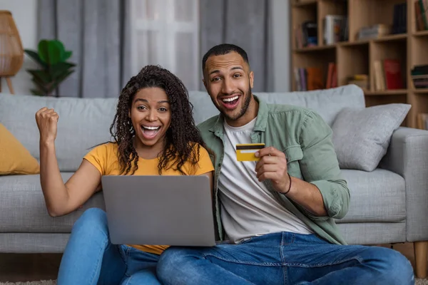 Emocionada pareja afroamericana con computadora portátil y tarjeta de crédito celebrando el éxito, reaccionando emocionalmente a los beneficios en línea — Foto de Stock