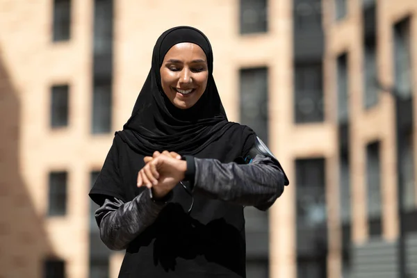 Perempuan arab muda yang ceria mengenakan jilbab dengan telepon di bahu memeriksa denyut nadinya pada pelacak kebugaran — Stok Foto