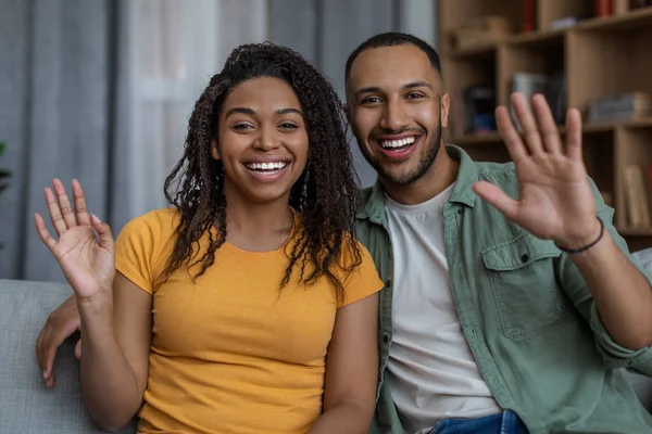 Cónyuges afroamericanos alegres saludando con las manos y sonriendo a la cámara, hablando en línea a través de videollamada, sentado en casa —  Fotos de Stock