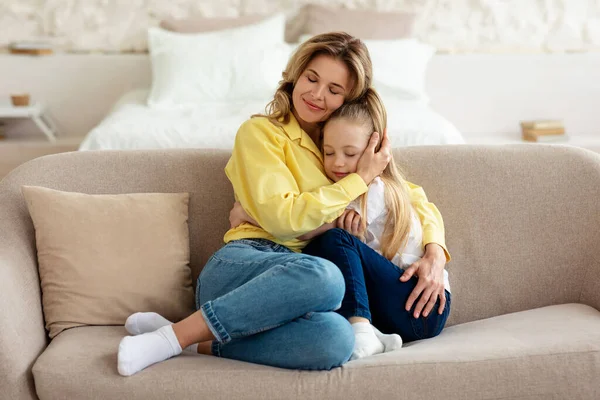 Älskade mamma kramar lilla dottern sitter på soffan hemma — Stockfoto