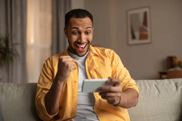 Joven hombre negro mirando el teléfono inteligente en la emoción, haciendo SÍ gesto en casa — Foto de Stock