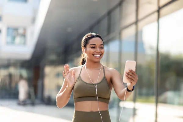 Atlético jovem senhora negra acenando na tela do smartphone, tendo videochamada online com treinador de fitness na rua da cidade — Fotografia de Stock