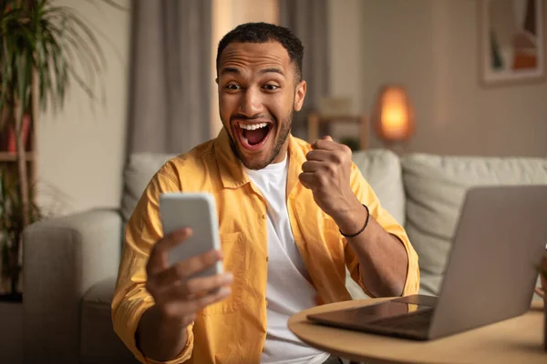 Захоплений молодий чорний чоловік дивиться на екран смартфона, жестикулює ТАК, відчуваючи себе щасливим через онлайн-перемогу вдома — стокове фото
