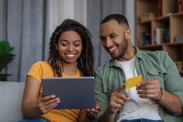 Cónyuges negros felices comprando en línea usando la tableta y la tarjeta de crédito, pagando la orden, sentados en el sofá en casa — Foto de Stock