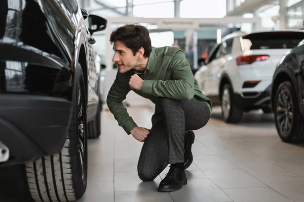 Pozytywny młody facet sprawdza koła nowego samochodu, kupuje nowoczesny pojazd w centrum sprzedaży samochodów — Zdjęcie stockowe