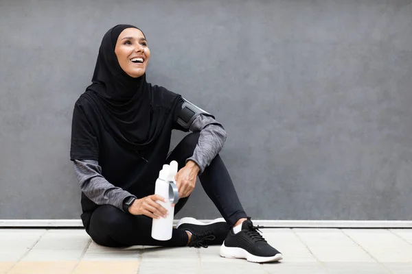 Wanita arab muda yang ceria dalam jilbab dengan pelacak kebugaran dan botol air duduk di lantai — Stok Foto