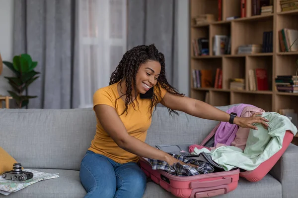 Σχέδιο ταξιδιωτικής προετοιμασίας. Ευτυχισμένη μαύρη γυναίκα πακετάρει βαλίτσα και ετοιμάζεται για διακοπές, κάθεται στον καναπέ στο σπίτι — Φωτογραφία Αρχείου