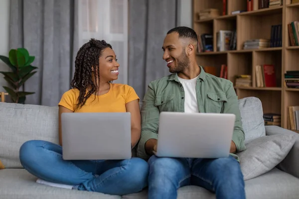 Unga svarta par använder bärbara datorer, tittar och ler mot varandra, sitter på soffan och surfar internet — Stockfoto