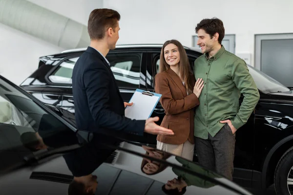 Jovem casal alegre conversando com vendedor de automóveis sobre compra de carro ou locação em concessionária moderna — Fotografia de Stock