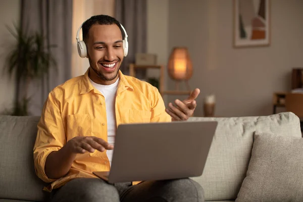 Joven hombre negro en los auriculares utilizando el ordenador portátil para el chat en línea, tener videoconferencia o reunión de negocios en casa — Foto de Stock