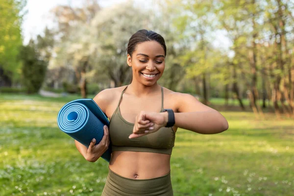 Happy black woman looking at smartwatch, tracking fitness activity, holding sports mat, ready for training outdoors — Stock Photo, Image