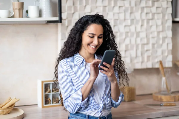 Retrato de una joven dama positiva usando un teléfono inteligente en la cocina, mensajería con amigos, pasar tiempo en casa — Foto de Stock