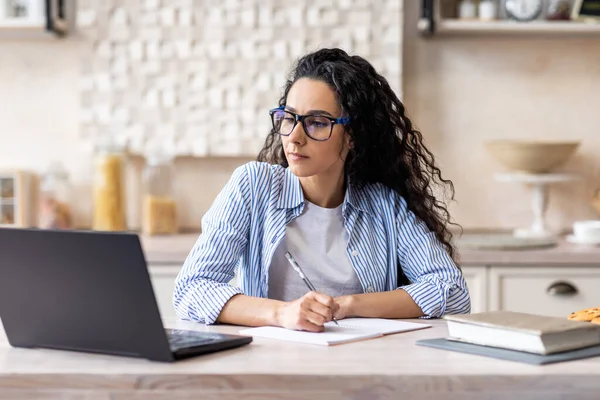 Jovem latina focada sentada na cozinha, tomando notas durante a aula on-line via laptop, espaço livre — Fotografia de Stock