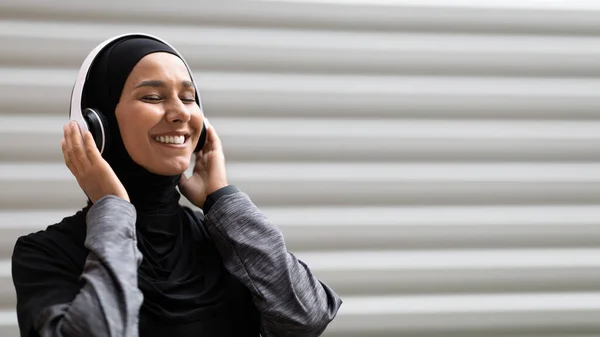 Happy young arab female in hijab in wireless headphone enjoy music on gray wall background outdoor — Stok Foto