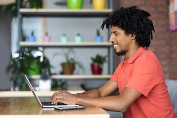 Feliz atraente encaracolado jovem preto masculino digitando no teclado do computador no interior do café com smartphone — Fotografia de Stock