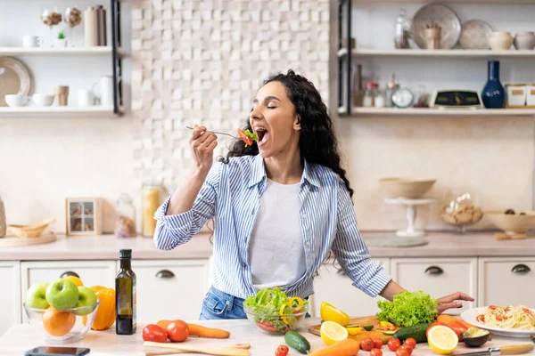 Молодая женщина ест салат за столом с органическими овощами, наслаждаясь здоровой диетой, стоя в светлом интерьере кухни — стоковое фото