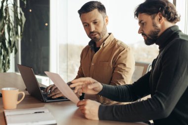 Ciddi şirket çalışanları belgeyi inceliyor, işbirliği yapıyor, modern ofiste dizüstü bilgisayar kullanıyor, boşluğu kopyalıyor