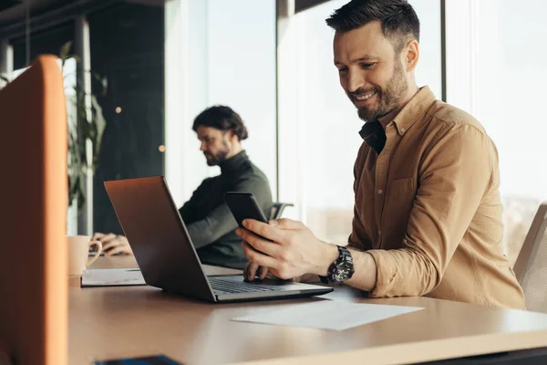 Glad manlig anställd anställd kontrollera smartphone, med hjälp av bärbar dator, arbeta med manlig kollega på moderna kontor — Stockfoto