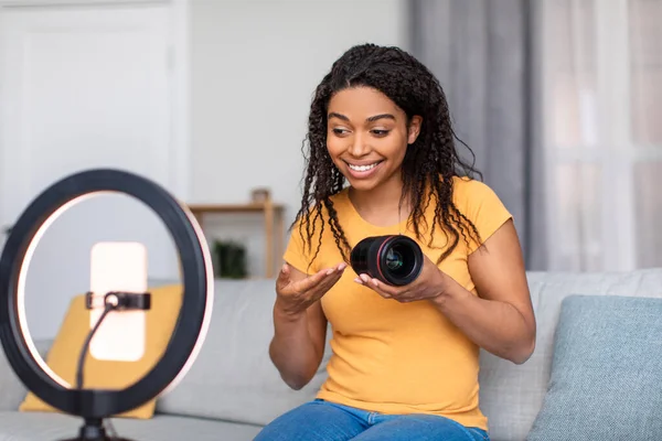 Happy african american female blogger recording review of camera lens, using cellphone and ring lamp