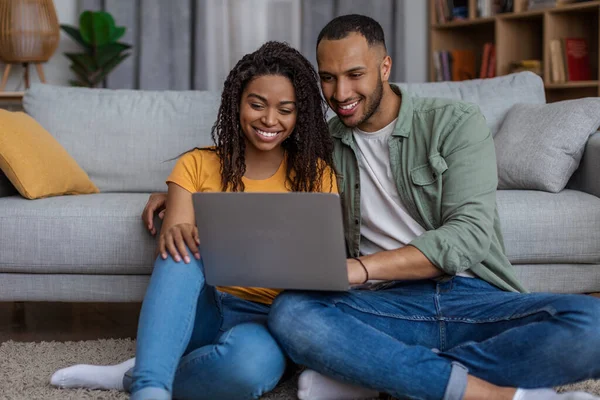 Pencinta Amerika yang positif afrika duduk di lantai oleh sofa dengan laptop, websurfing dan bersenang-senang bersama di rumah — Stok Foto