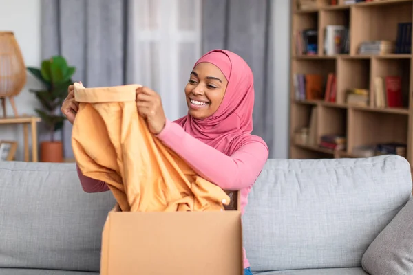 Happy black muslim woman opening carton box, checking her delivery, holding new clothes and smiling — Stock Photo, Image