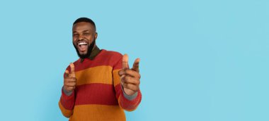 Yakaladım seni. İki eliyle kameraya poz veren neşeli, yakışıklı siyahi adam, Milenyum Afrikalı Amerikalı erkek, birini işaret ediyor ve gülümsüyor, mavi stüdyoda eğleniyor,