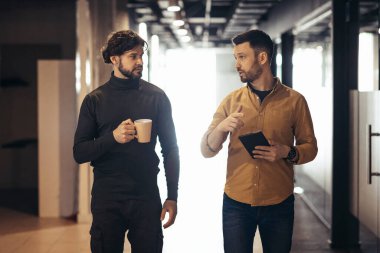 Erkek iş arkadaşları iş projesini tartışıyor, ofiste kahve molası sırasında şirket salonunda yürürken tablet bilgisayar kullanıyorlar.