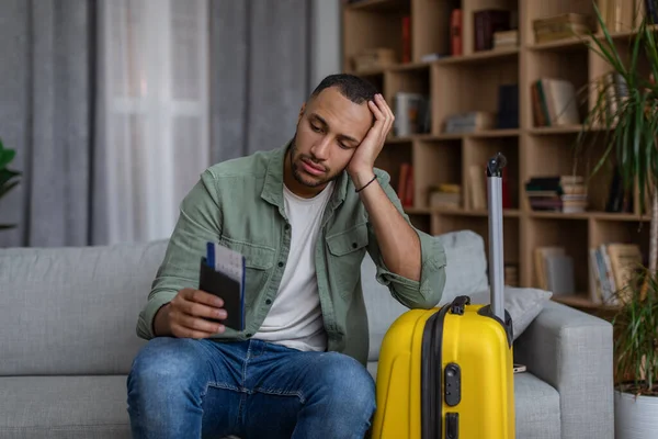 Homme afro-américain bouleversé avec passeport et billets d'avion assis sur le canapé près de valise emballée, a manqué ses vacances — Photo
