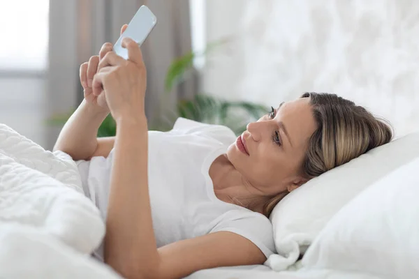 Pozitivní dáma ležící v posteli sama, pomocí mobilního telefonu — Stock fotografie