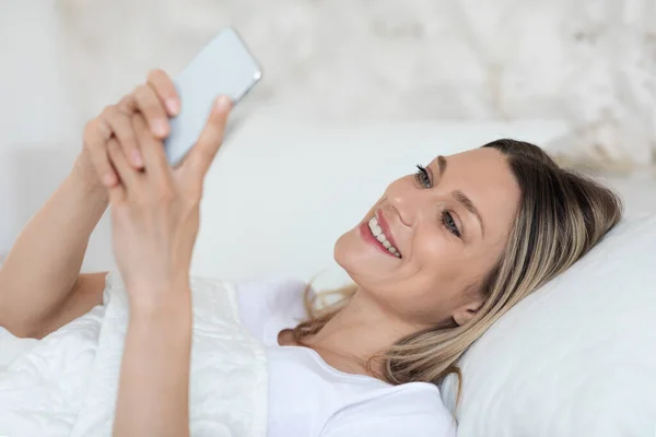 Весела блондинка відпочиває в ліжку з мобільним телефоном — стокове фото