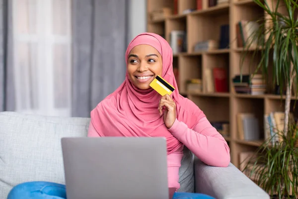 Bolti mániás. Álmodozó fekete muszlim hölgy kezében hitelkártya és a laptop, gondolkodás vásárlások interneten keresztül — Stock Fotó