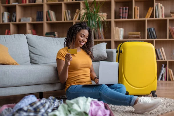 Senhora preta alegre com cartão de crédito reserva férias na agência de viagens, usando laptop e planejamento viagem ao exterior em casa — Fotografia de Stock