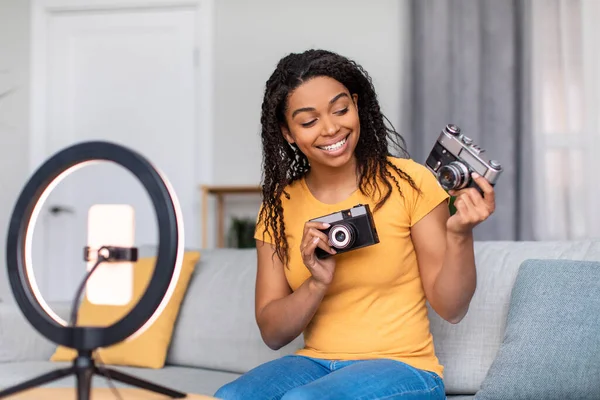 Heyecanlı zenci kadın blogger evdeki kanepede otururken iki eski kameranın video incelemesini kaydediyor — Stok fotoğraf