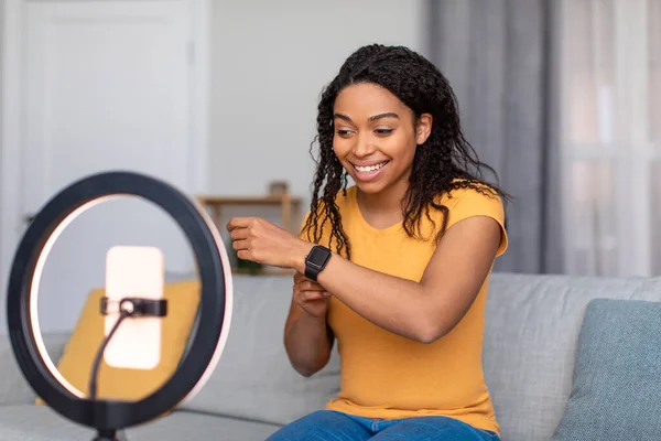 Konsep blogging. Happy african american female video blogger with cellphone on ring lamp recording review on smartwatch — Stok Foto