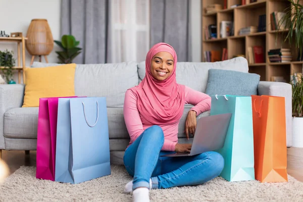 Bilgisayarlı, alışveriş çantalı, internet üzerinden alışveriş yapan, yerde oturan ve gülümseyen Müslüman bir kadın. — Stok fotoğraf