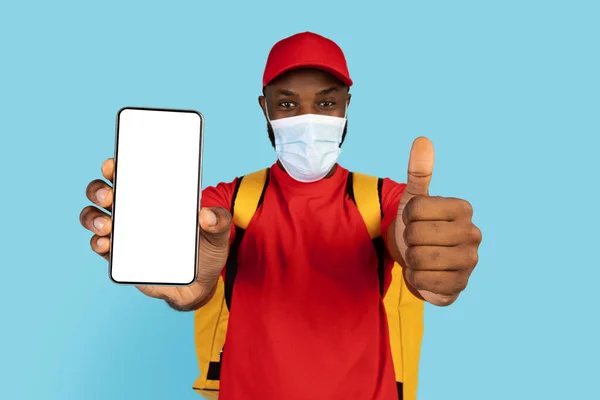 Schwarzer Lieferheld mit medizinischer Maske zeigt leeres Smartphone und Daumen hoch — Stockfoto