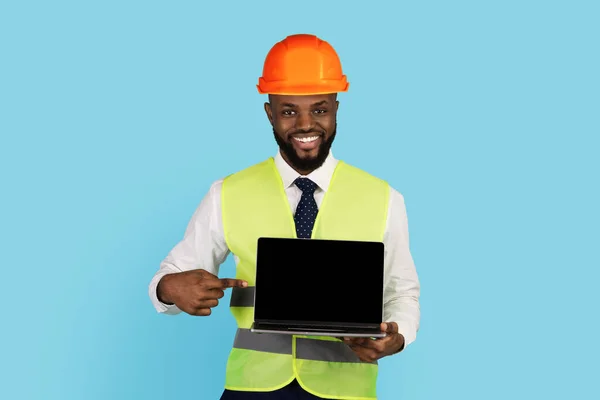 Sorrindo preto trabalhador de construção apontando para laptop com tela em branco na mão — Fotografia de Stock