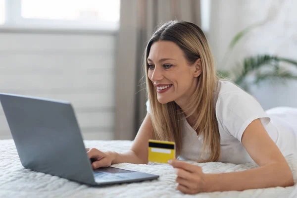 Mujer joven feliz con ordenador portátil y tarjeta de crédito en el dormitorio — Foto de Stock