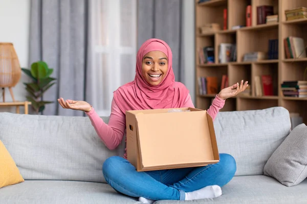 Overjoyed african american muslim lady opening cardboard box, receiving online shop order at home, sitting on sofa