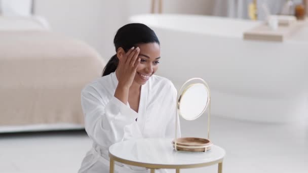 Jovem senhora afro-americana feliz vestindo roupão acariciando seu rosto, desfrutando da condição de sua pele, sorrindo ao espelho — Vídeo de Stock