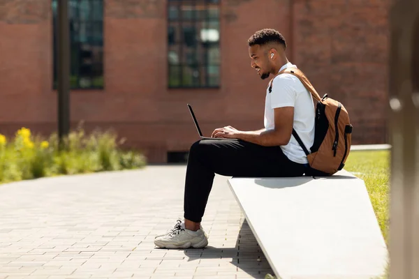 Schwarzer Mann mit drahtlosen Kopfhörern benutzt Laptop im Freien — Stockfoto