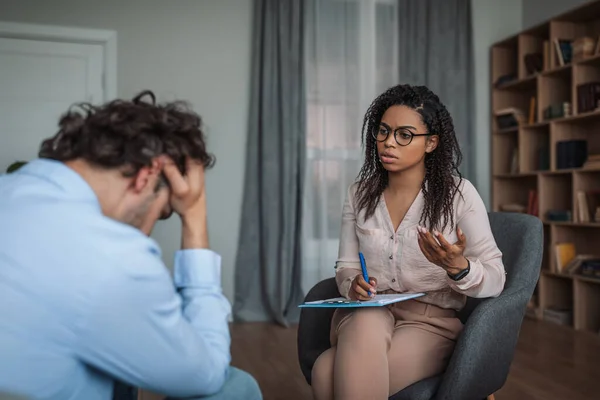 Ernstige jonge Afrikaanse Amerikaanse dame psycholoog consulting huilen depressieve Europese man in kliniek — Stockfoto