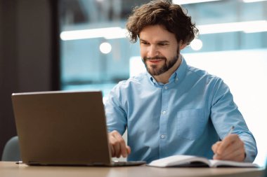 Dizüstü bilgisayar kullanan ve not defterine yazan gülümseyen bir adamın portresi