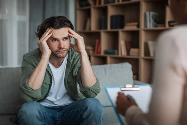 Verdrietig jong europees man op overleg met Afrikaanse amerikaanse dame psycholoog in moderne kliniek interieur — Stockfoto