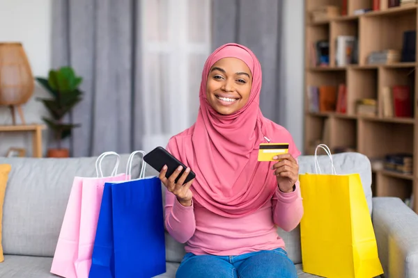 Alışveriş torbaları, kredi kartı ve akıllı telefon ile evden alışveriş yapan güzel siyah Müslüman kadın, koltukta oturuyor. — Stok fotoğraf