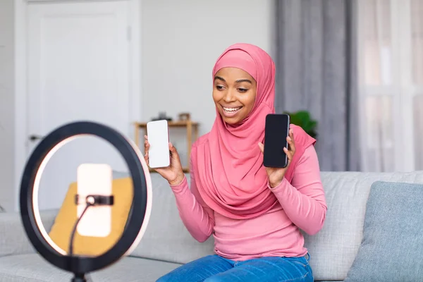 Cheerful muslim black blogger pemfilman video review dari dua smartphone baru, berbicara dengan kamera web ponsel pada lampu cincin — Stok Foto