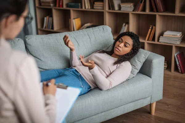 Sério jovem afro-americano paciente feminino encontra-se no sofá, consulta com dama médica europeia — Fotografia de Stock