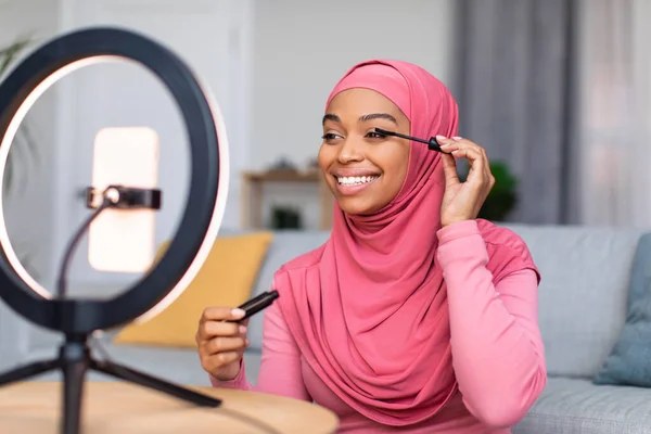 Pelajaran tata rias online dari blogger kecantikan. Wanita kulit hitam yang senang mengenakan maskara pada bulu mata, melihat smartphone — Stok Foto