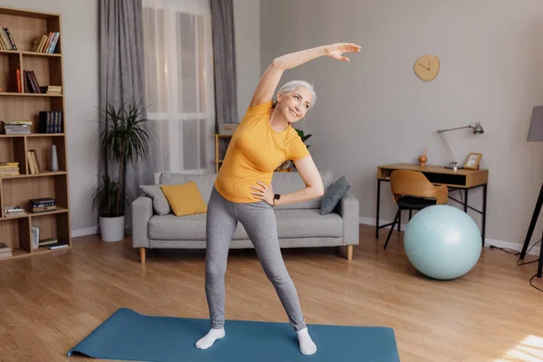 Спорт на пенсии. Счастливая пожилая женщина делает упражнения гибкости для здорового образа жизни во время домашней тренировки — стоковое фото